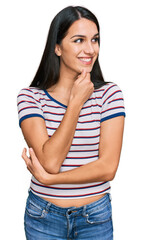 Young hispanic girl wearing casual striped t shirt with hand on chin thinking about question, pensive expression. smiling with thoughtful face. doubt concept.