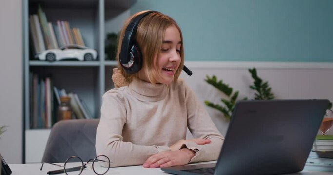 Beautiful little Caucasian girl in headphones wave hand greeting with tutor, study online lesson with teacher using laptop. Homeschooling concept