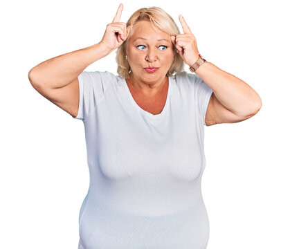 Middle age blonde woman wearing casual white t shirt doing funny gesture with finger over head as bull horns