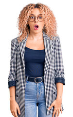 Young blonde woman with curly hair wearing business jacket and glasses afraid and shocked with surprise and amazed expression, fear and excited face.