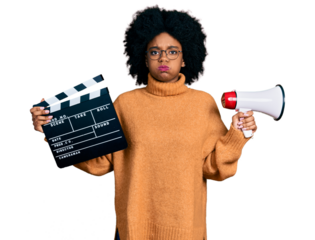 Young african american woman holding video film clapboard and megaphone puffing cheeks with funny face. mouth inflated with air, catching air.