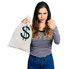 Beautiful hispanic woman holding dollars bag annoyed and frustrated shouting with anger, yelling...