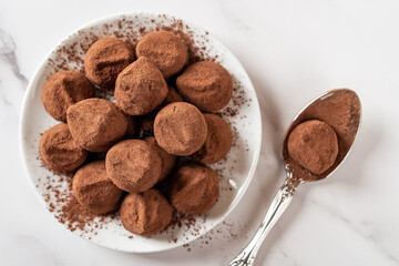Gourmet chocolate truffles on a white saucer. Plate full of dark chocolate candy balls in cocoa powder and spoon over marble surface. Sweet food, dessert and confection concept. Macro.