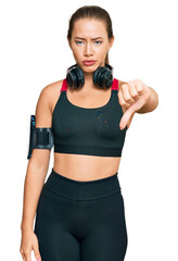 Beautiful blonde woman wearing gym clothes and using headphones looking unhappy and angry showing rejection and negative with thumbs down gesture. bad expression.