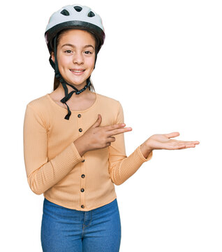 Beautiful brunette little girl wearing bike helmet amazed and smiling to the camera while presenting with hand and pointing with finger.