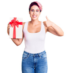 Young beautiful woman with pink hair holding gift smiling happy and positive, thumb up doing excellent and approval sign