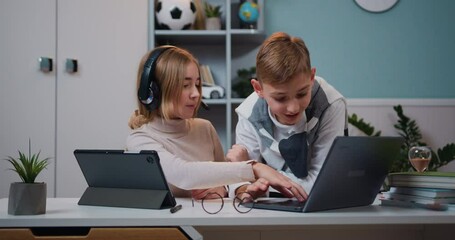 Small cute girl with headphones using internet games on digital tablet while kid boy playing game on laptop in living room at home, family concept - Powered by Adobe