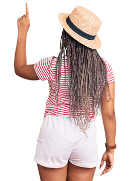 Young african american woman with braids wearing summer hat posing backwards pointing ahead with finger hand