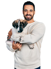 Young hispanic man with beard hugging cute dog smiling with a happy and cool smile on face. showing...