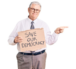 Senior grey-haired man wearing business clothes holding save our democracy protest banner smiling happy pointing with hand and finger to the side