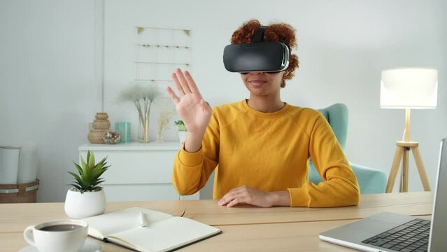 African american young woman wearing using virtual reality metaverse VR glasses headset at home. Girl touching air during VR experience on virtual reality helmet. Simulation hi-tech videogame concept