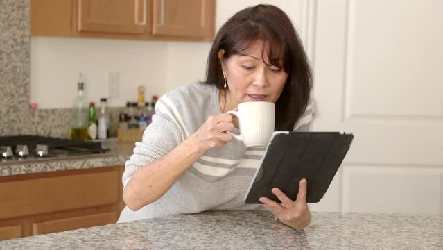 Attractive Stylish Woman In Grey Sweater Leaning On Bright Kitchen Counter In Peaceful Home Smiling Enjoying Reading IPad Mobile Tablet Drinking Coffee. Beautiful Asian Female Has Cheerful Lifestyle.