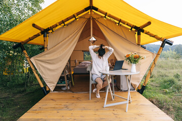 young woman working on a laptop while sitting near a big tent in nature