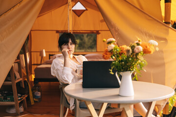 Pretty woman using cellphones and notebook sitting in tent during camping on summer day
