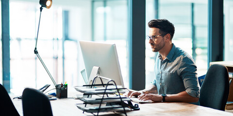 The successful stay consistent. Shot of a focused young businessman using a computer at his desk in...