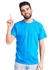 Young handsome man with beard wearing casual t-shirt pointing finger up with successful idea. exited and happy. number one.