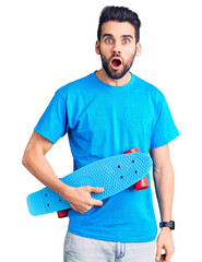 Young handsome man with beard holding skate scared and amazed with open mouth for surprise,...