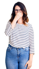 Fototapeta premium Beautiful young brunette woman wearing casual clothes and glasses smelling something stinky and disgusting, intolerable smell, holding breath with fingers on nose. bad smell