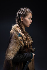 A young brave Viking heroine with a sword in hands on a dark background. A young woman in a suit in...
