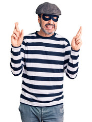 Naklejka premium Middle age handsome man wearing burglar mask gesturing finger crossed smiling with hope and eyes closed. luck and superstitious concept.