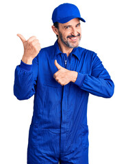 Middle age handsome man wearing mechanic uniform pointing to the back behind with hand and thumbs...