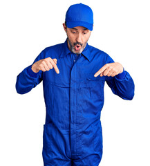 Middle age handsome man wearing mechanic uniform pointing down with fingers showing advertisement, surprised face and open mouth