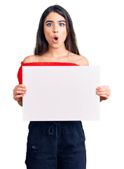 Brunette teenager girl holding blank empty banner scared and amazed with open mouth for surprise, disbelief face