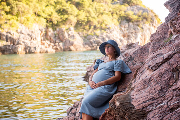 Middle aged pregnant woman in blue dress, hat sits on rock by the sea. Millennial girl travels in pregnancy on vacation. Family holiday on the ocean. Natural texture. Merging with nature. Copy space