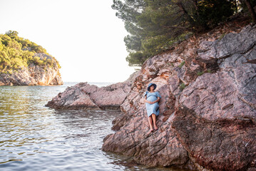 Middle aged pregnant woman in blue dress, hat sits on rock by the sea. Millennial girl travels in pregnancy on vacation. Family holiday on the ocean. Natural texture. Merging with nature. Copy space