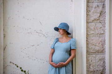 Obraz na płótnie Canvas Portrait of middle aged pregnant woman in denim blue dress, hat. Future mother stands near antique ruins, grunge background green ivy. Travel during pregnancy. Maternity, pregnancy concept. Copy space