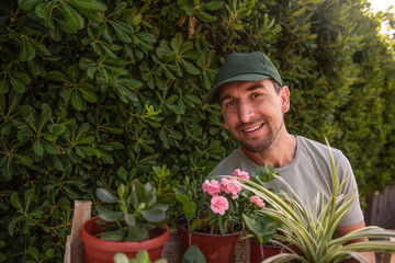 Man gardener in green cap smiles as smells indoor plants in front of living evergreen fence Phillyrea latifolia. Allergy to flowers. Natural holiday gift. Small business, hobby, gardening. Copy space