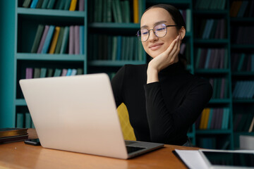 A female freelancer smiling with glasses using a laptop on a desk is in a modern office. Formal wear for young people.