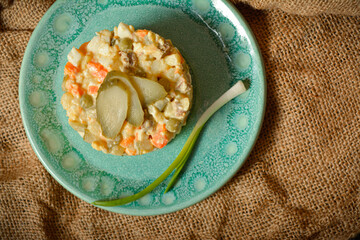 Directly above of traditional rustic salad Olivier served in culinary ring. Country Salad with potato, carrot, peas, pickled cucumbers, eggs on sackcloth textile background. Spring onion near dish