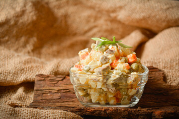 Close-up to Traditional Russian salad Olivier served in glass bowl on tree bark. Salad with potato and carrot cubes, green peas, pickled cucumbers, eggs on sackcloth textile background
