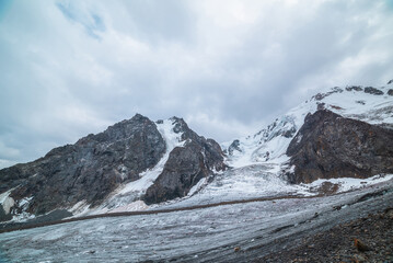 Dramatic landscape with glacier tongue and icefall on large snow mountain range with sharp rocky pinnacle under gray cloudy sky. Long glacier in high altitude. Gloomy scenery in mountains in overcast.