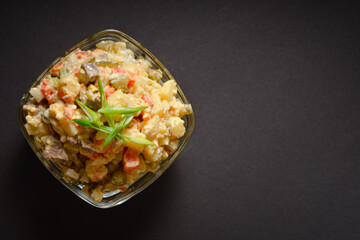 Top view of Traditional Russian salad Olivier served in glass bowl. Salad with potato and carrot cubes, green peas, pickled cucumbers, eggs on Dark background