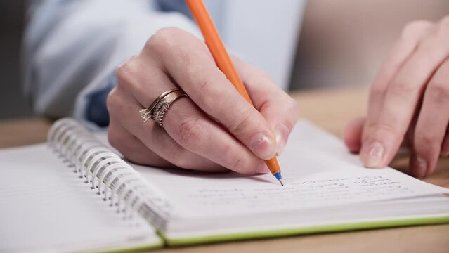 young woman works, holds a pen in her hands and makes notes in a notebook while sitting at a table, close-up