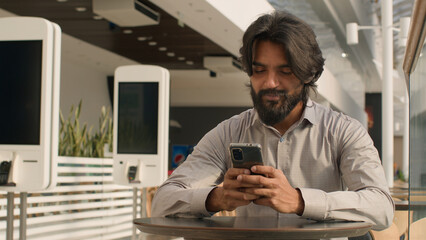 Happy young indian businessman holding smartphone sending message sitting indoors smiling arabian...