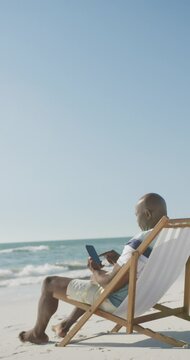 Vertical video of senior african american man using smartphone on deckchair at beach, in slow motion
