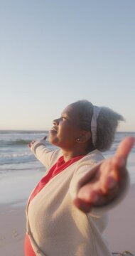 Vertical video of happy senior african american woman widening arms at beach, in slow motion