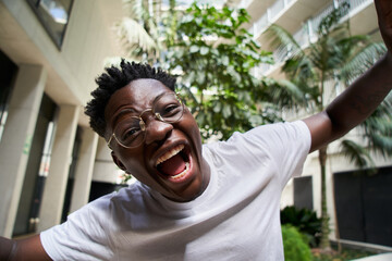 Selfie with cell of a young African American man with his mouth open screaming with happiness. Smiling people looking at camera outside. Portrait of extremely excited black boy. Close up people. 
