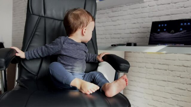 Little baby boss sitting comfortably in a big leather chair. Cute boy takes tv remote from a table.
