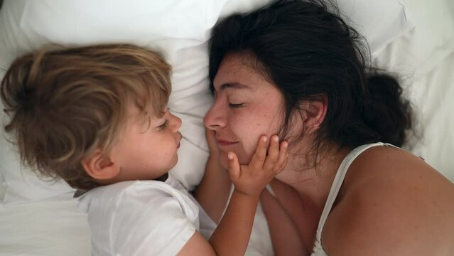 Cute moment between mother and child laying in bed in the morning. Adorable small boy holding mom face. Family lifestyle affection and care