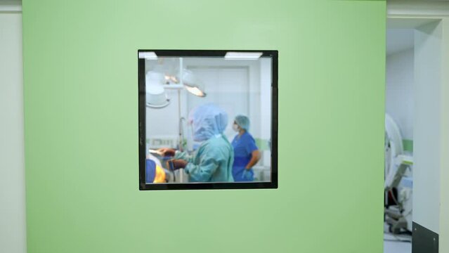 Square window in a door to a light modern surgery room. Doctor in protective suit applies iodine on the patient. Female nurse leaves the room.
