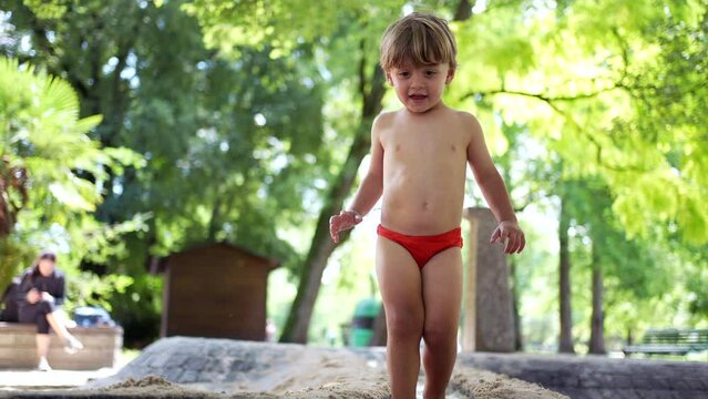 Joyful small boy wearing red bathing suit briefs jumping in the air splashing into park puddle water in slow motion. Happiness childhood lifestyle happiness
