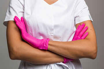 A woman in a white lab coat and pink latex gloves stands with her arms crossed against a light background. Medical staff