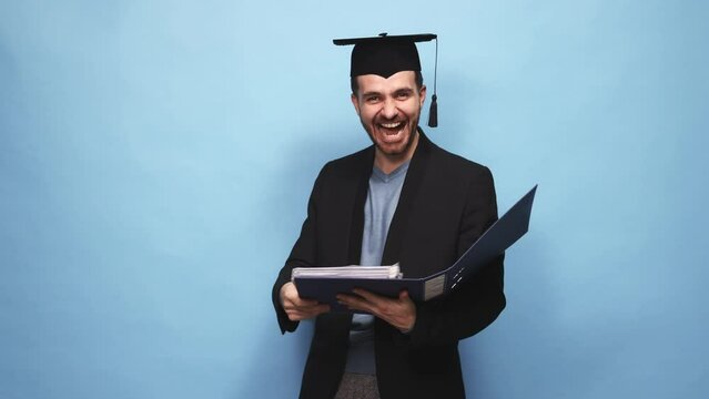Graduation. Happy and excited young man, student in graduation costume, getting diploma against blue studio background. Concept of education, success, personal growth, achievements, education