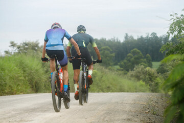 Em um dia ensolarado, ciclistas vestidos com equipamento apropriado para a prática esportiva se aventuram em uma estrada rural de terra, apreciando a paisagem natural composta por árvores verdes.