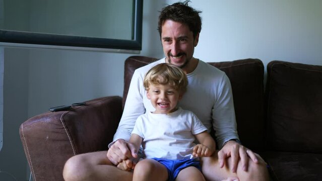 Playful dad and son relationship. Child seated on father lap on couch at home. Authentic real life fatherhood lifestyle