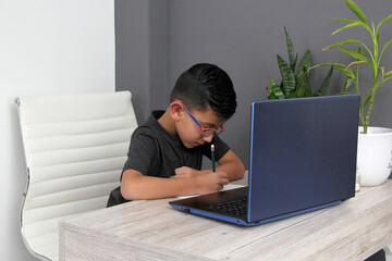 7-year-old Latino boy with glasses does home schooling takes online classes at home on a desk with a laptop, studies, is surprised and participates in class